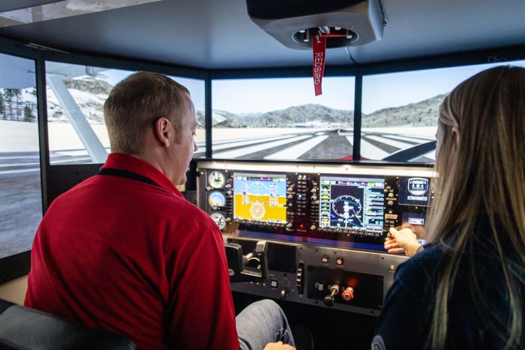 An Aviation instructor guides a student through a flight simulator lesson while reviewing the cockpit displays and controls.