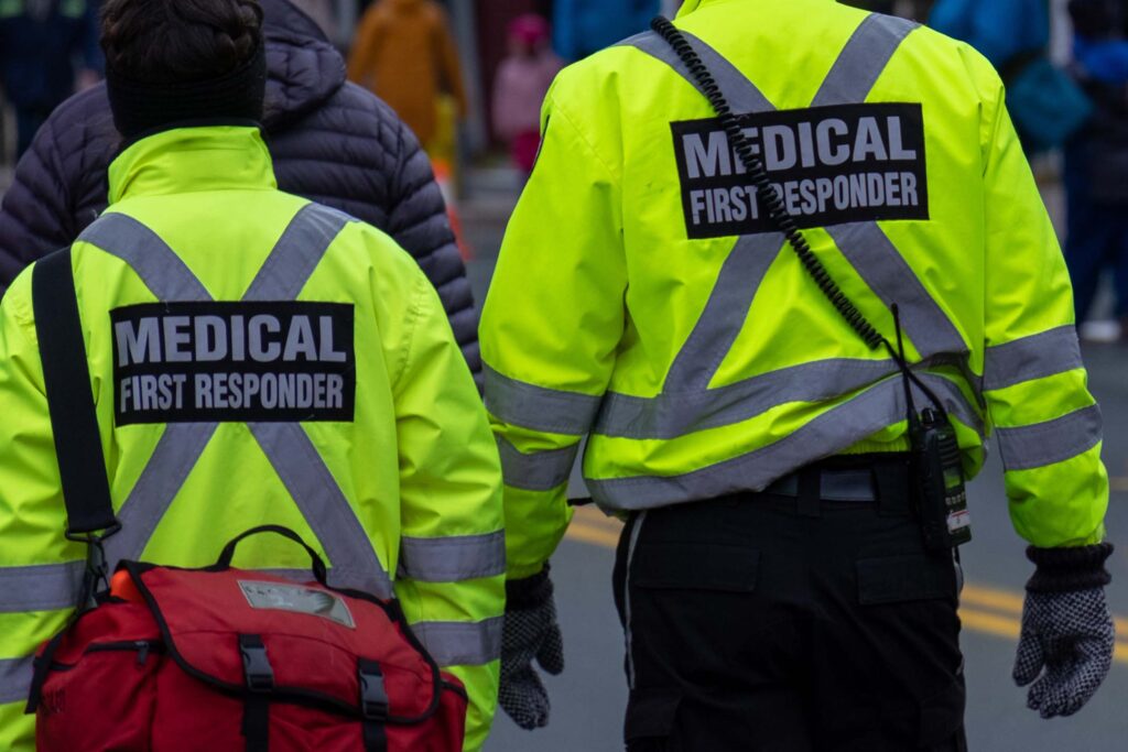 Two Advanced Emergency Medical Technicians in high-visibility jackets walking away from the camera at an outdoor event.