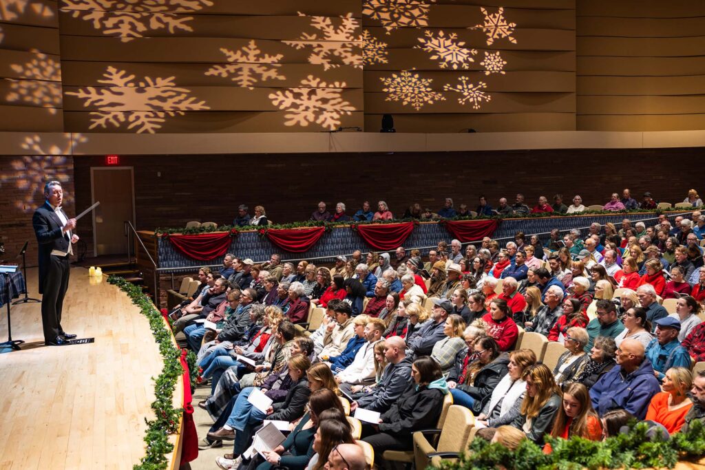Conductor leads an orchestra on stage as an audience watches in a decorated concert hall.