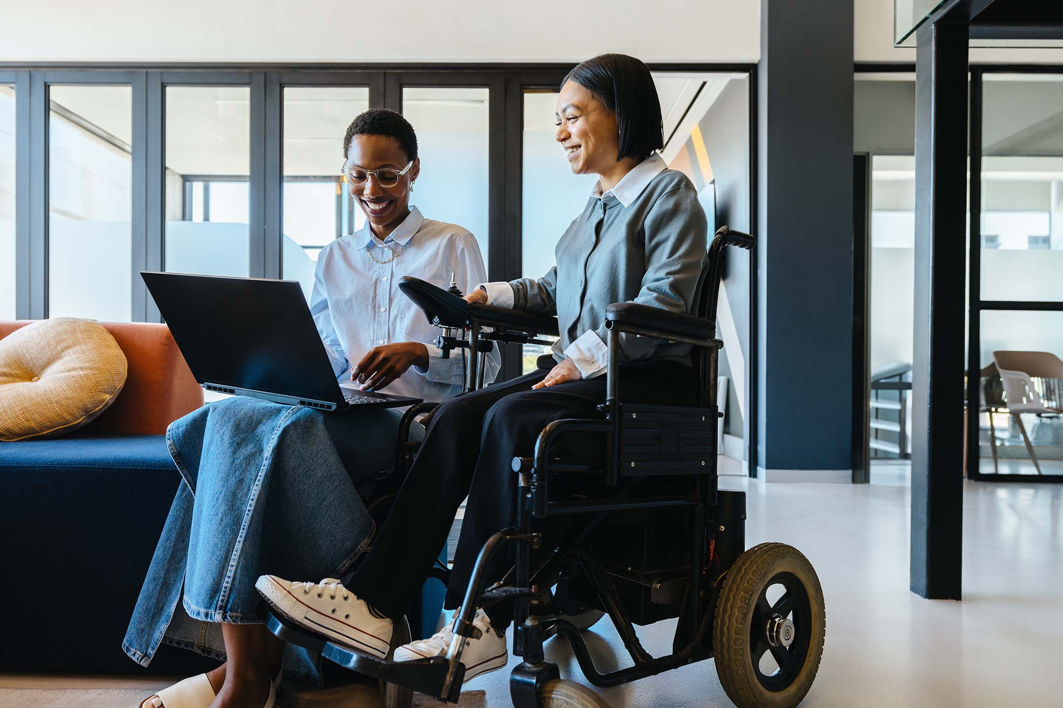 Two women in a modern office, one using a wheelchair, working together with a laptop as she uses her assistive technology degree.
