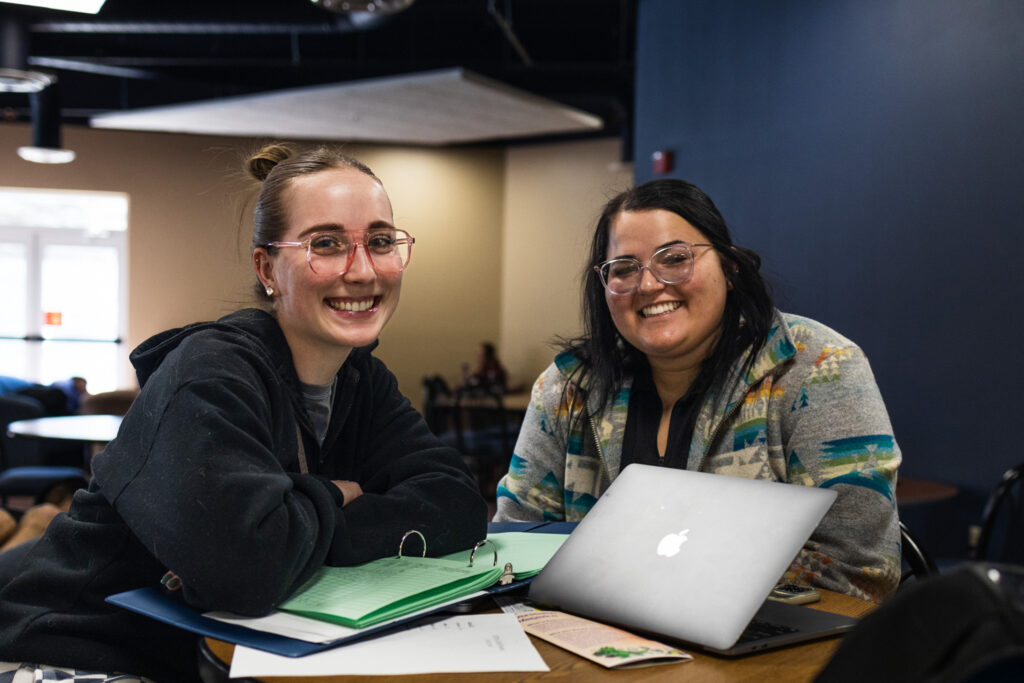 Two students sit together at a table with a laptop and papers, smiling while studying in a campus common area.
