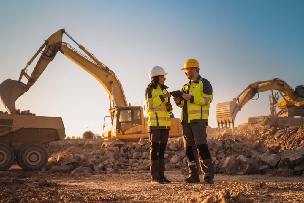 Two construction workers in hard hats standing at a job site with heavy equipment behind them getting taught construction tech.