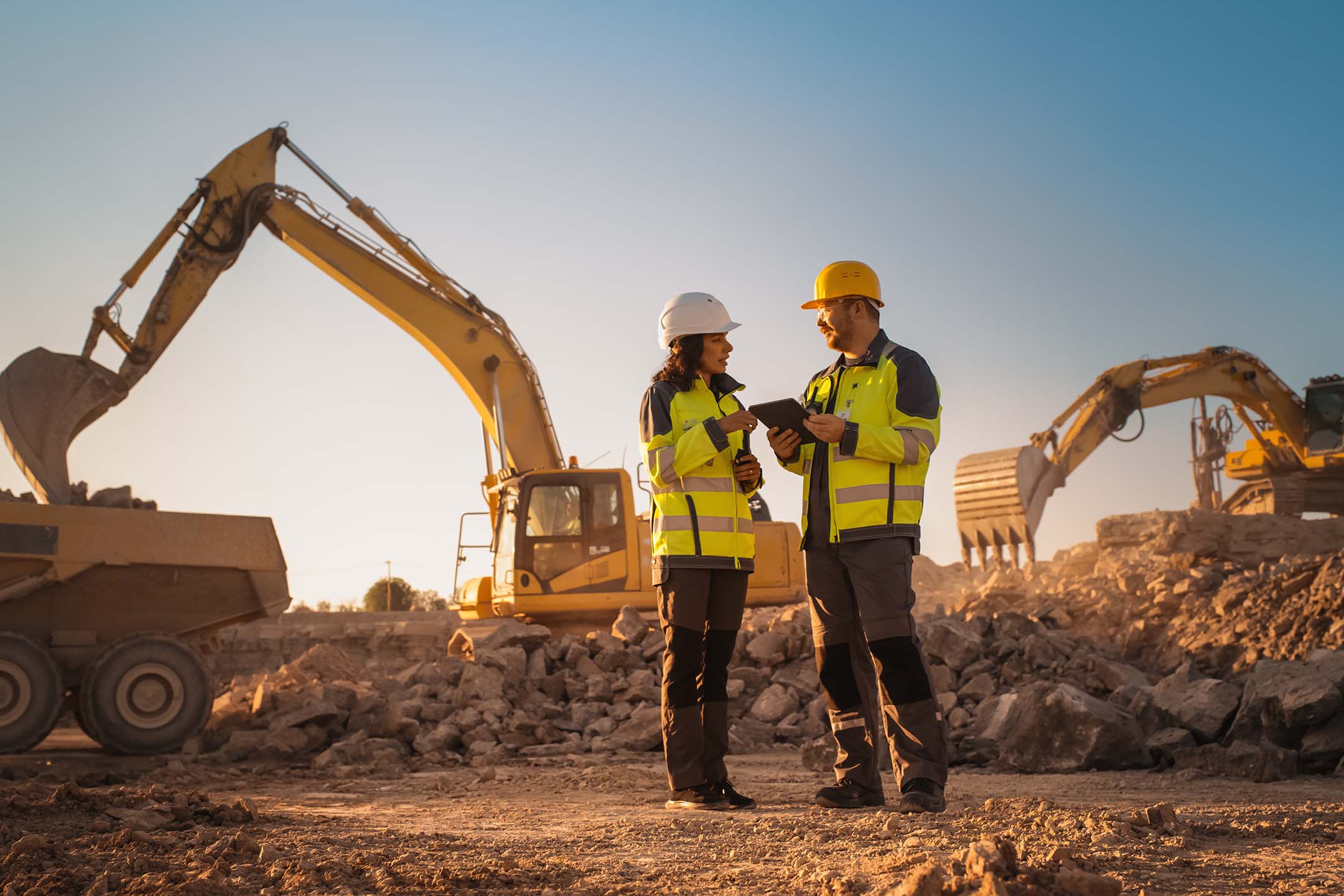 Two construction workers in hard hats standing at a job site with heavy equipment behind them getting taught construction tech.