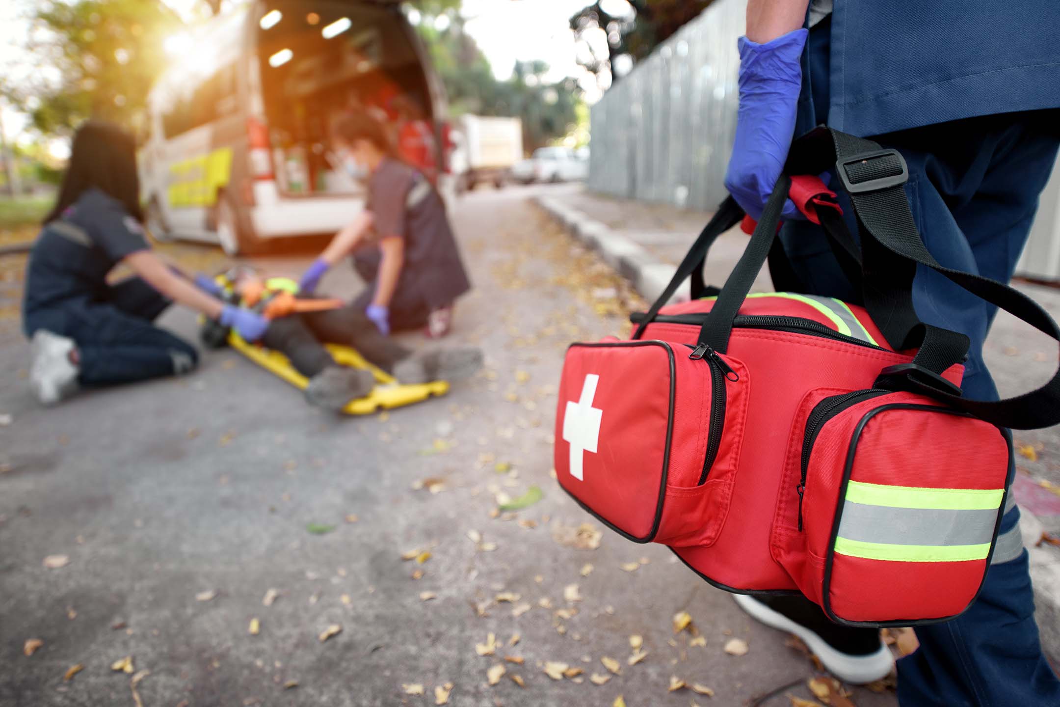 First aid bag in the foreground with emergency responders assisting someone in the background who learned paramedic technology.