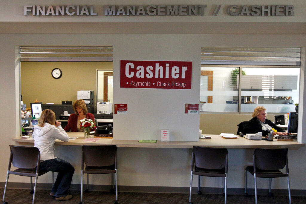 Students seated at a service counter labeled ‘Cashier,’ speaking with staff members behind glass windows.
