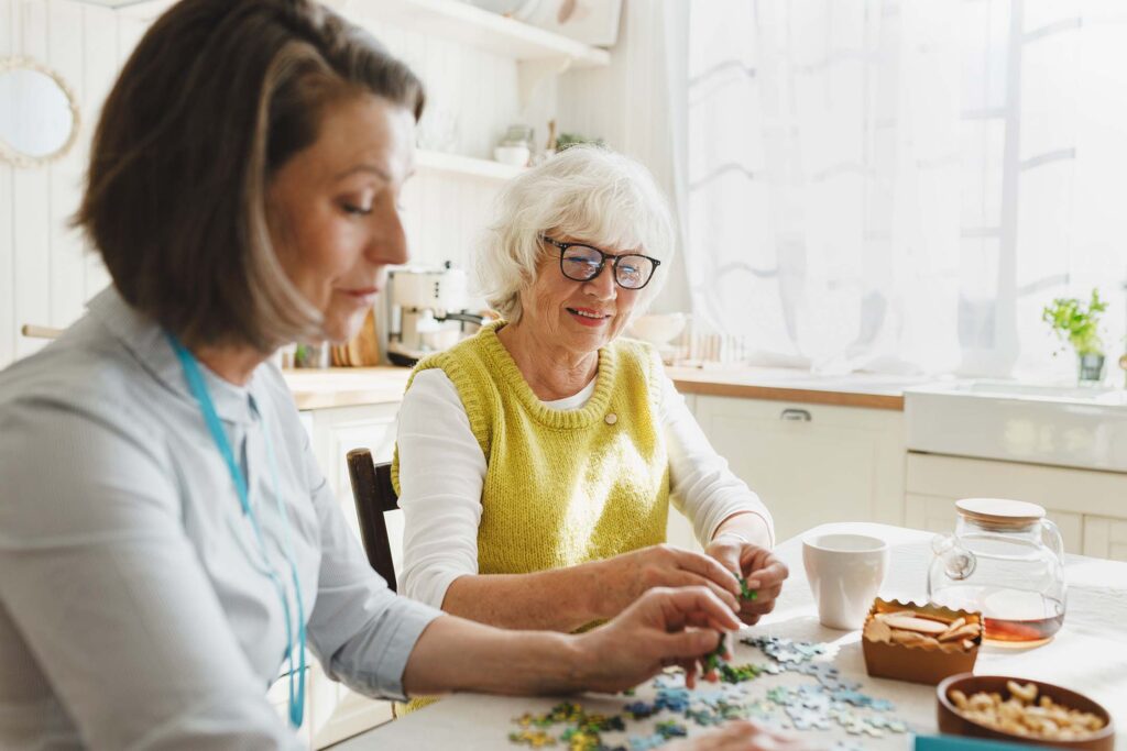 Caregiver helping an older woman with a craft or hands-on activity at a table. This is gerontology.
