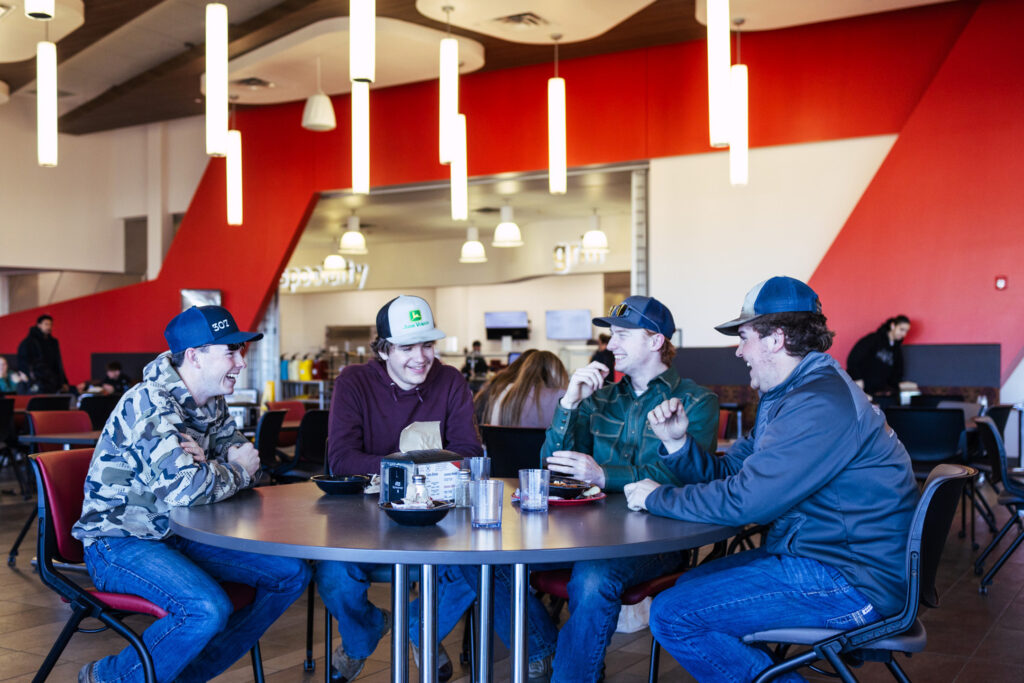 Group of students seated around a table in a campus dining area, laughing and talking while eating together.