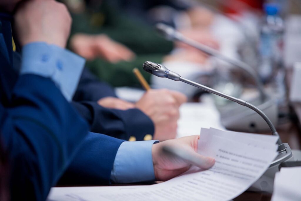 People seated at microphones and documents during a formal meeting or panel discussion. Political science