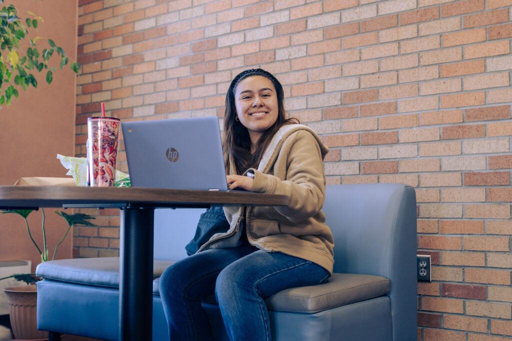 Student seated at a table using a laptop in a campus lounge with brick walls and natural light.