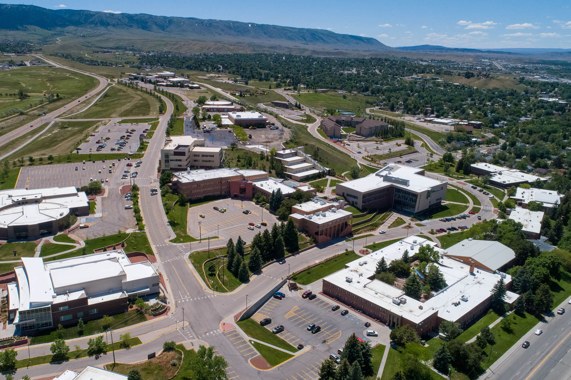 High-angle aerial drone view of the Casper College campus in Wyoming, showing various academic buildings, parking lots, and green spaces against a backdrop of rolling hills and a clear blue sky.