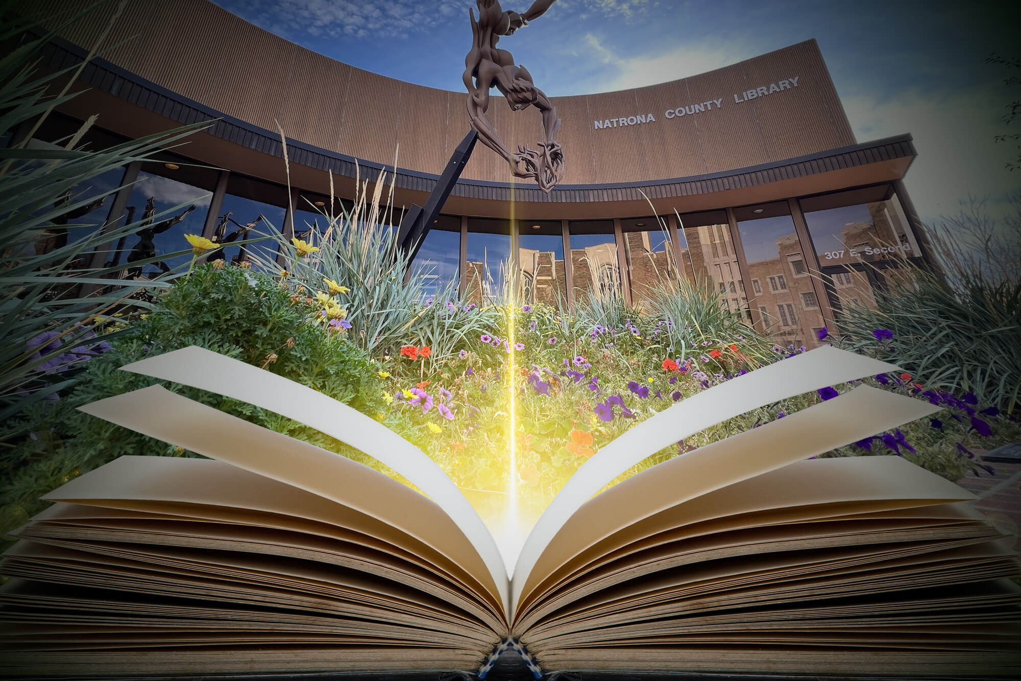 An open book with glowing light emanating from its pages in front of the Natrona County Library building and its "The Oracle" sculpture.