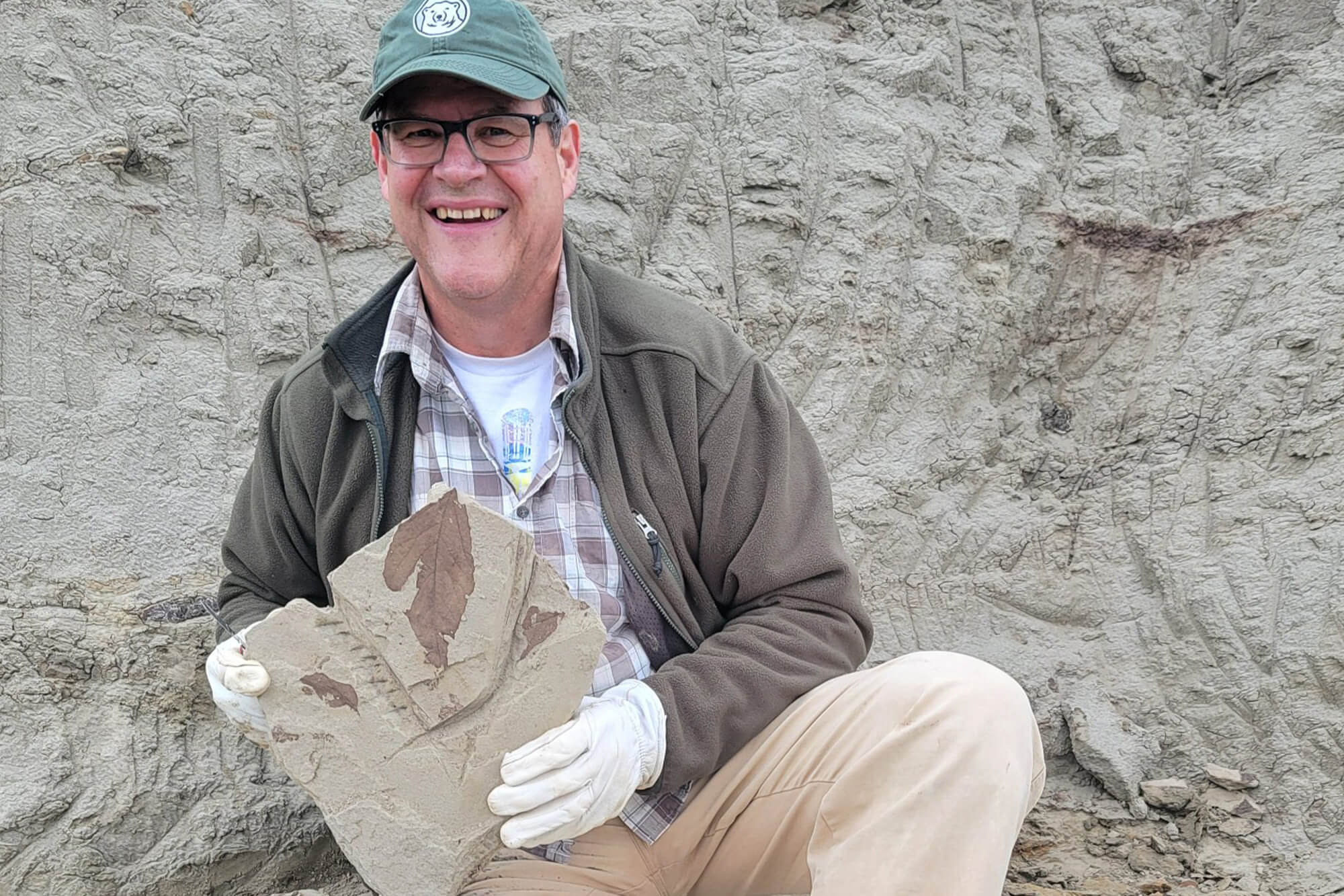 Kirk Johnson, Ph.D., kneeling outside and holding a stone slab containing a clear prehistoric leaf fossil specimen.