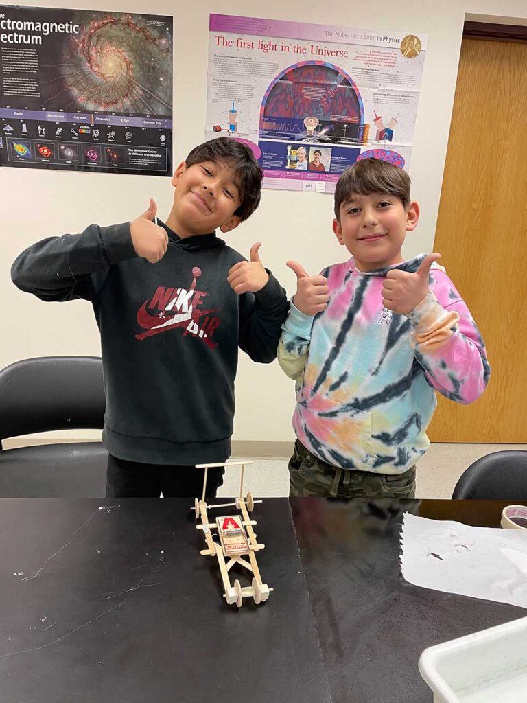 Two smiling students give a thumbs up behind a small, handmade mousetrap car on a classroom table