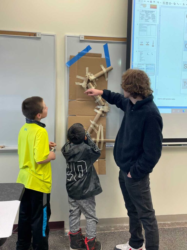 An instructor points to a cardboard marble run taped to a whiteboard while two young students look on during a classroom activity