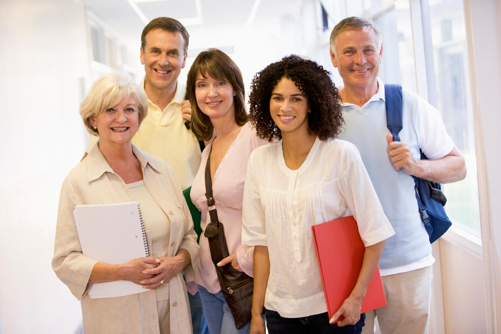 Five smiling adult community education students standing in a brightly lit hallway holding notebooks and folders.