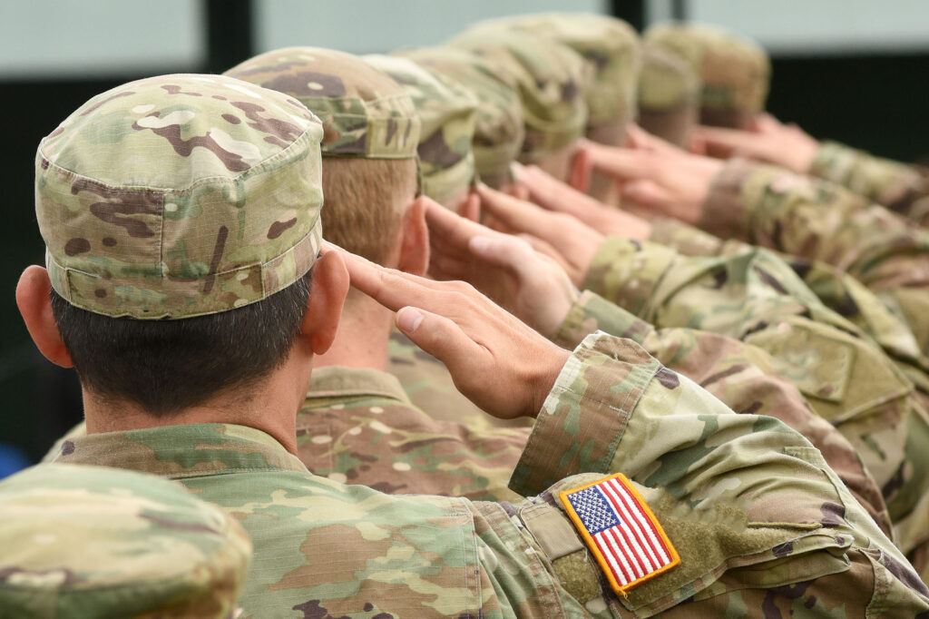 Row of U.S. military service members in camouflage uniforms saluting with the American flag patch visible on the shoulder.