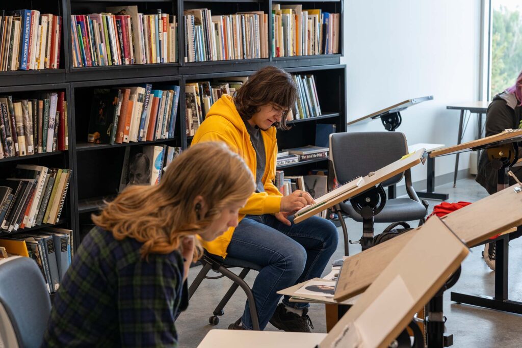 Art students work on drawing assignments at easels in a bright studio classroom.