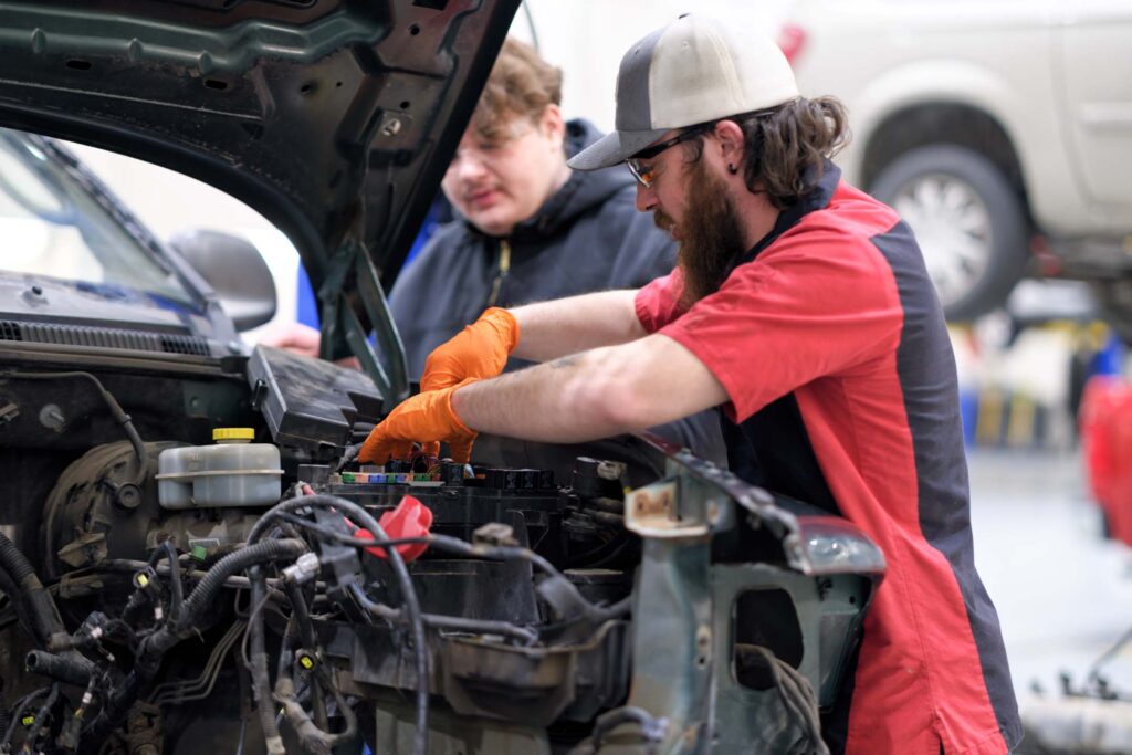 An Automotive Technology student wearing safety glasses and gloves works under the hood of a vehicle while another student observes.