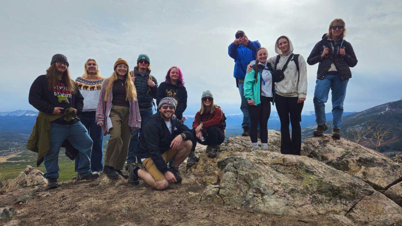 A group of Casper College students pose together on a rocky mountain overlook with snow-capped mountains and a cloudy sky in the background.