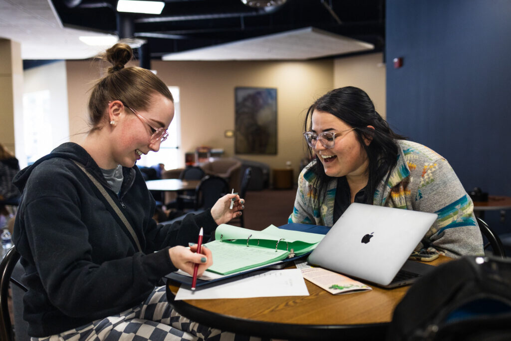 Two students smiling and reviewing coursework together at a table with a laptop and open binder as they choose a major.