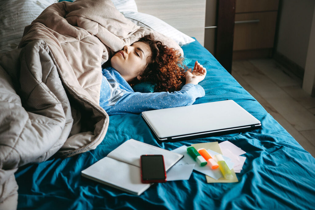 Student resting in bed surrounded by schoolwork and electronics illustrating habits tied to college success.