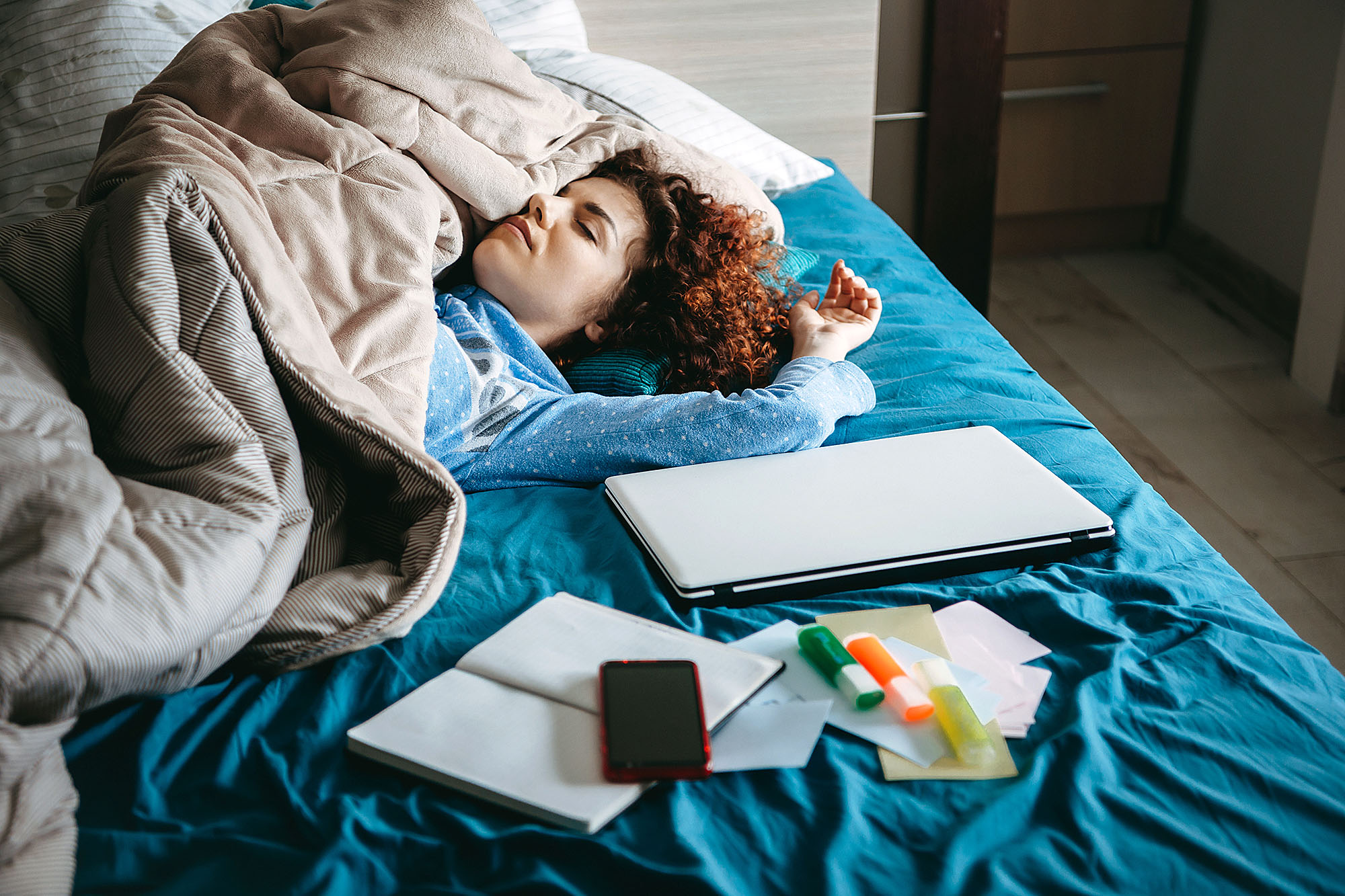 Student resting in bed surrounded by schoolwork and electronics illustrating habits tied to college success.